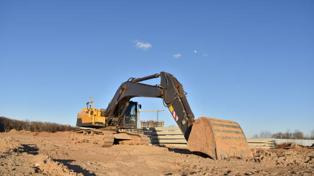 Excavator on earthworks at construction site. Backhoe on foundation work and road construction. Tower cranes in action on blue sky background. Heavy machinery and construction equipment